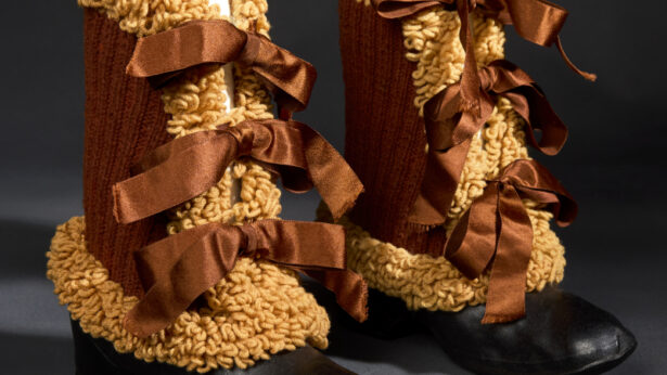 Two boots with shiny black rubber bottoms and brown knitted yarn shanks adorned with curly looped yarn and three brown, satin ribbon closures siting side by side.