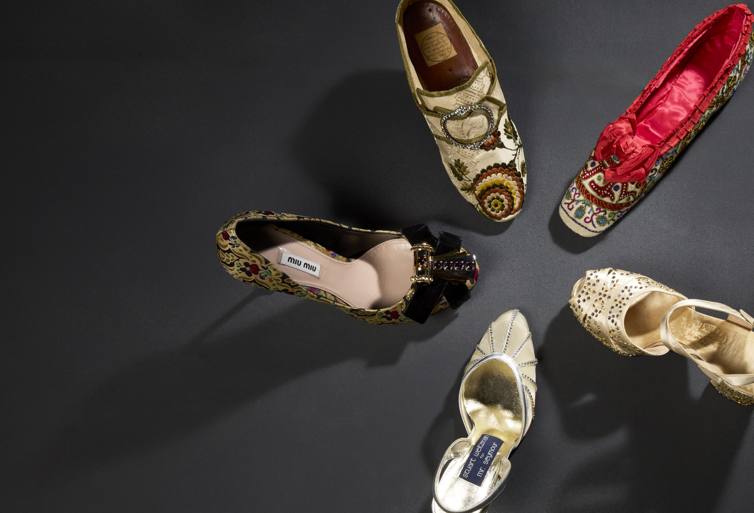 A circle of five women's shoes. Clockwise: eighteenth century, English silk brocade slipper with buckle closure, 1860s embroidered slipper with red silk lining, 1940s I. Miller platform sandal with metallic embellishment, 1980s Stuart Weitzman sling-back heel, 2010s Miu Miu brocade pump with velvet bow and shoe clip.