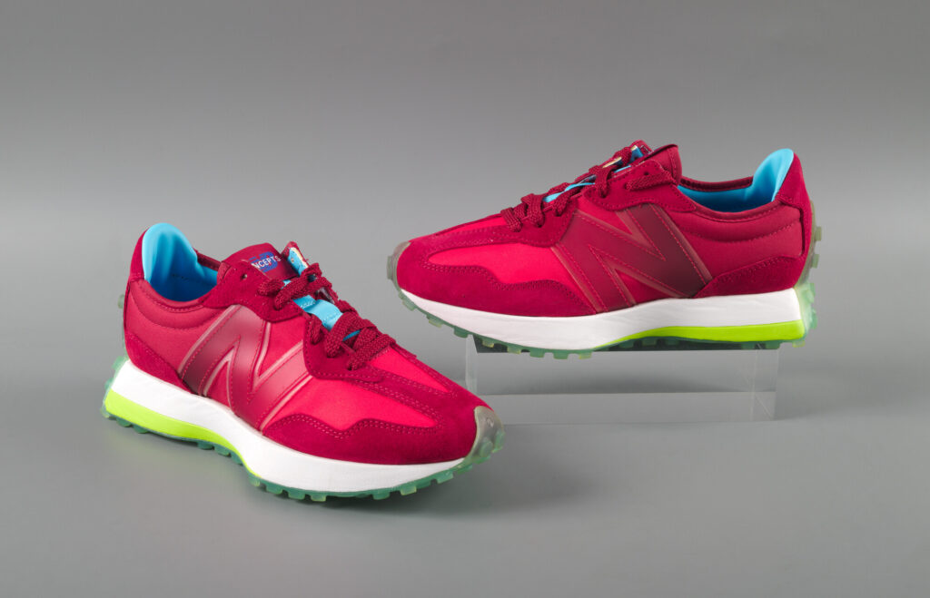 A pair of multi-tone red suede and synthetic fabric athletic sneakers with large “N” on side, bright blue interior, and white foam soles with bright green at bottom, and clear light blue tread.