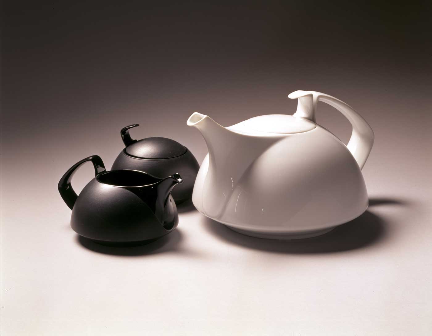 An angular, modernist white teapot next to two smaller black teapots of similar style.