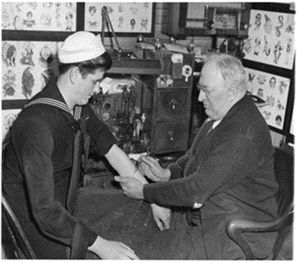 A black and white photograph of a sailor receiving a tattoo from an artist, with both seated in front of panels advertising tattoo designs.