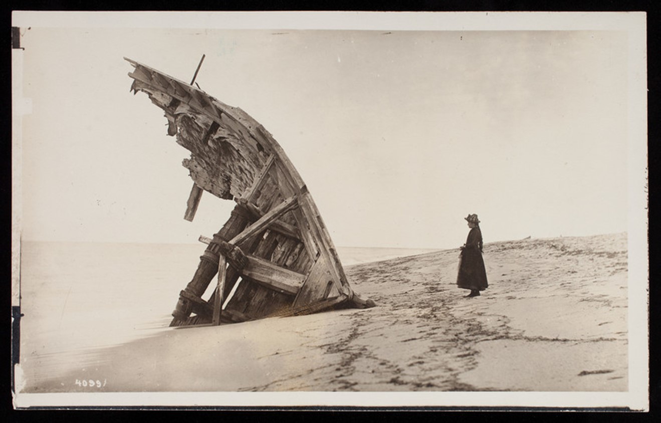 ID: A black and white photograph of a shipwreck. A woman in dark dress and light hat with braided hair looks at the remains of the bow of the ship on the edge of a beach.