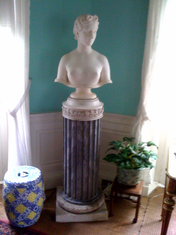 ID: A photograph of the full statue of the Bust of Psyche. It is a marble statue depicting the bare bust of a slender woman with soft features and ringlets in her hair. A black pillar supports the sculpture in a room with a blue wall, a patterned blue and yellow vase to the left, and a green plant to the right.