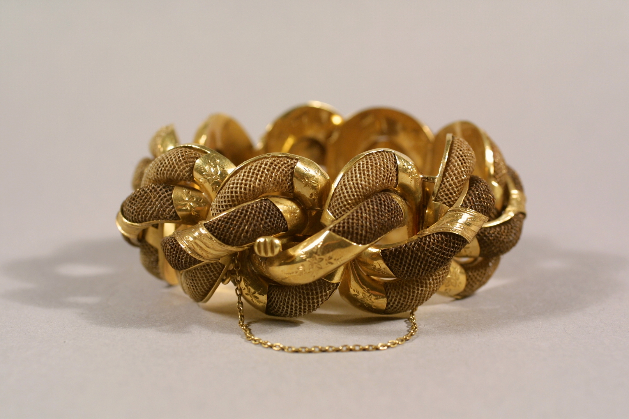 ID: A golden hair bracelet weaved with interlocking segments of gold chain and sold gold with a smaller gold chain attached and hanging underneath