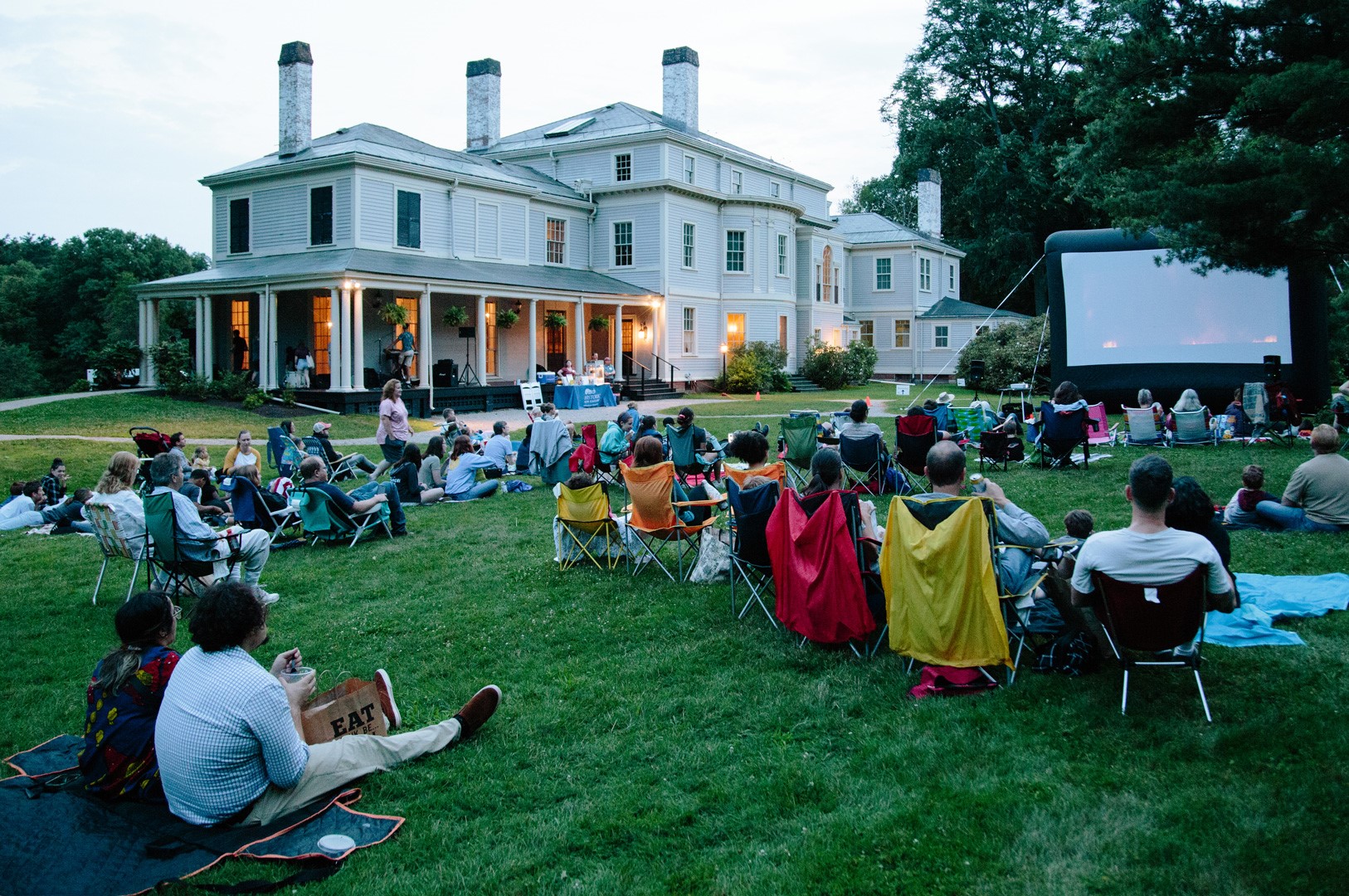A large group of people sitting on lawn chairs and blankets on the rear lawn of the Lyman Estate, with an inflatable movie screen in front of them. It is early evening, and warm lights are visible through the windows of the mansion behind them.