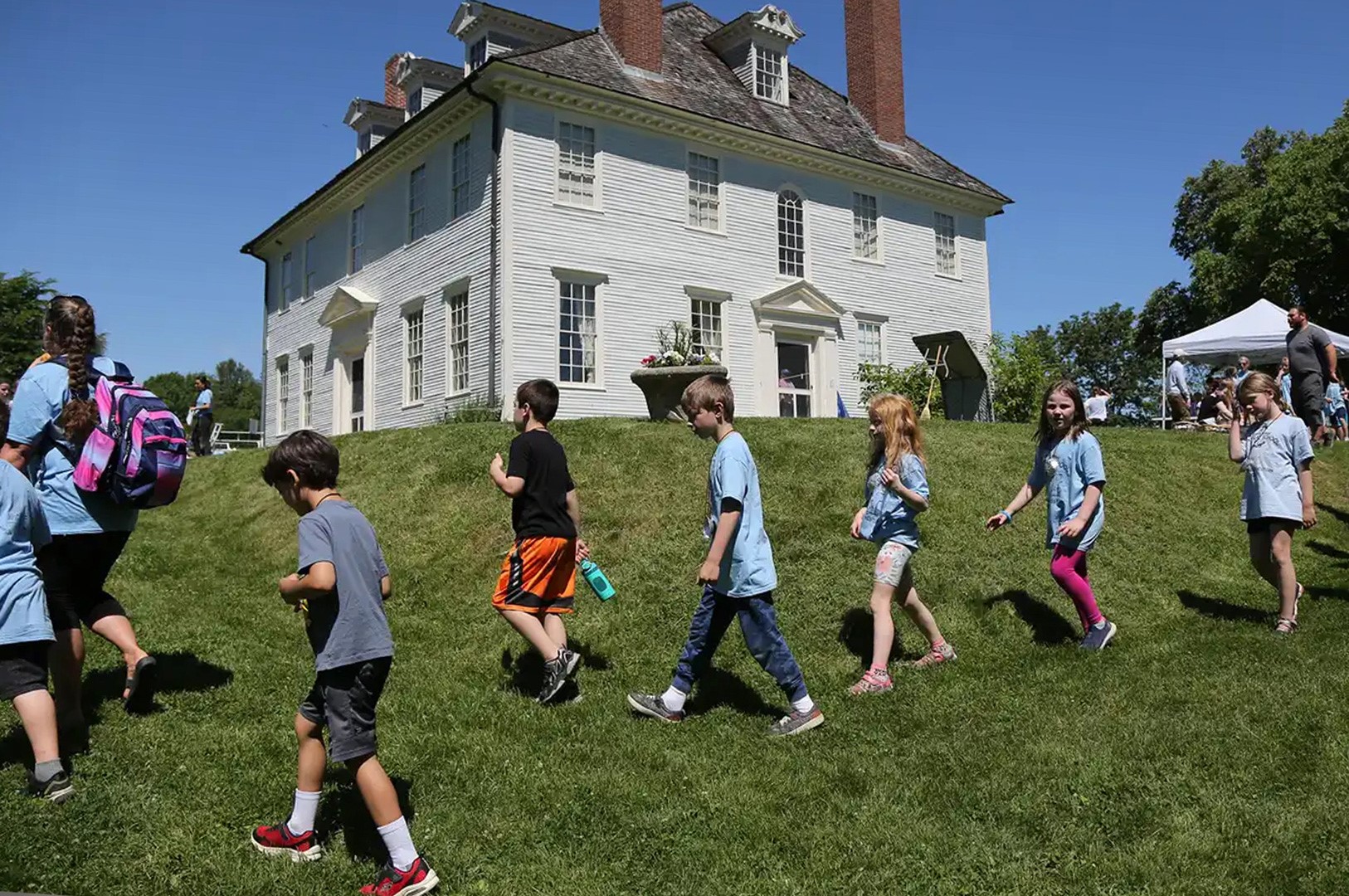 A line of children following an adult with a back pack from a pop-up tent across the lawn of a large historic house.