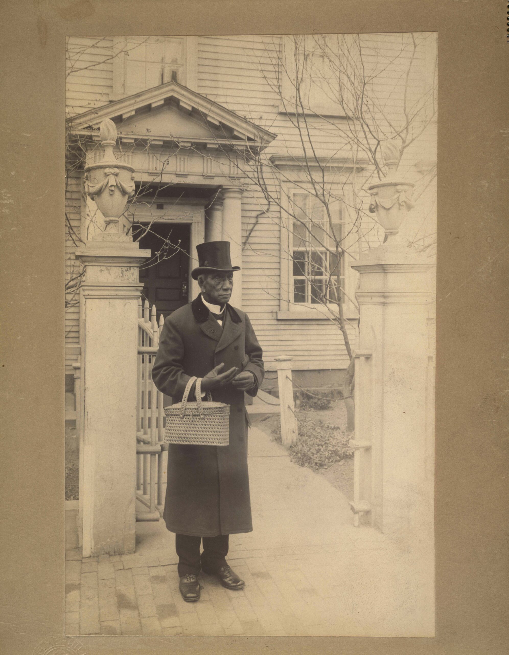 Black and white photograph of an African American man wearing a trench coat and tophat carrying a basket in front of a gate to a home